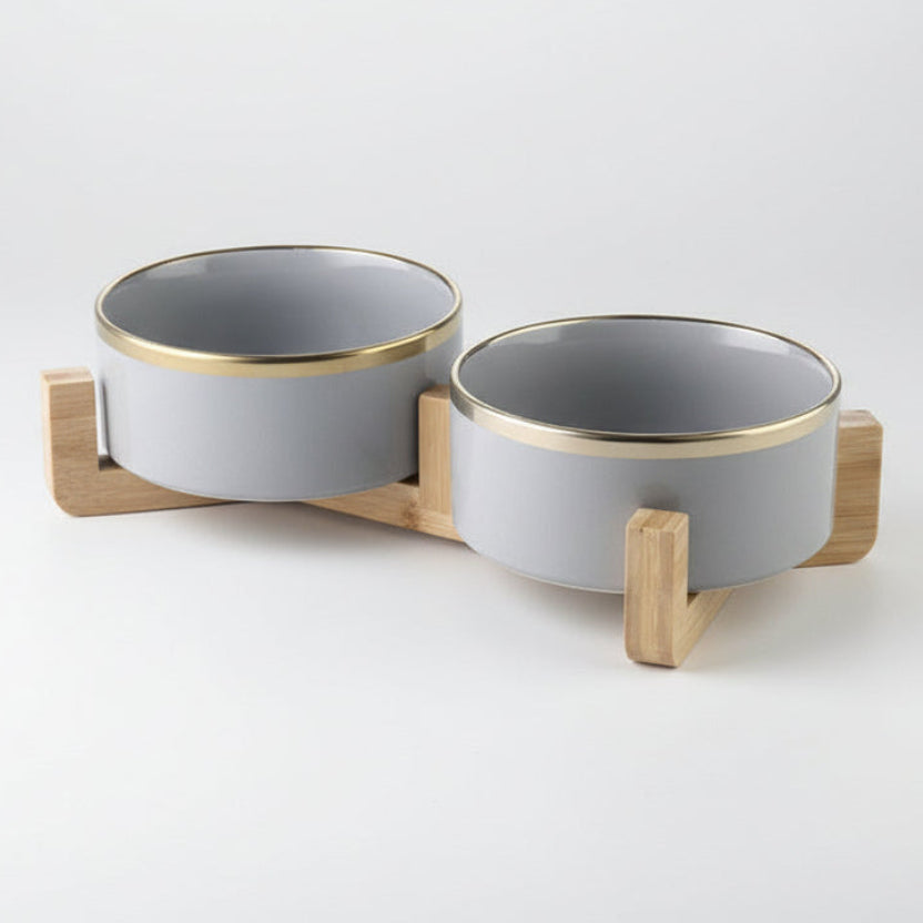 ​Ensemble de deux gamelles haut de gamme pour chat en céramique blanche brillante avec liserés dorés élégants, surélevées par un support minimaliste en bois de chêne clair, signée Palais Félin Paris.