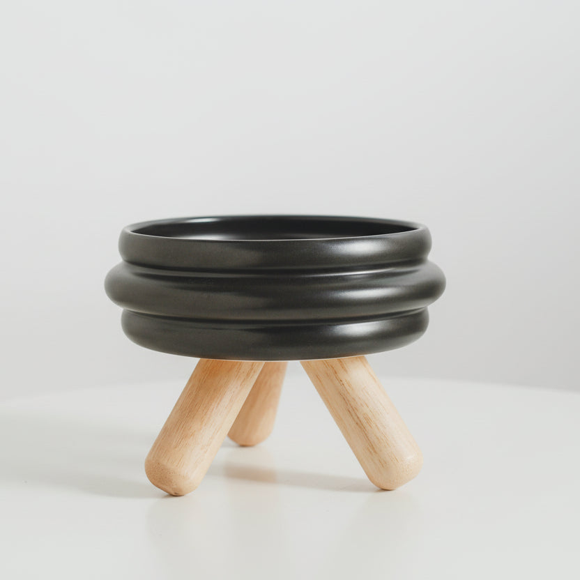 ​Gamelle individuelle au design moderne pour chat en céramique noire mate, posée sur un support minimaliste en bois de chêne clair croisé, isolée sur un fond blanc, signée Palais Félin Paris.