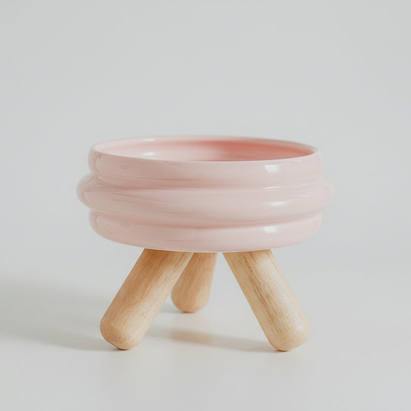 ​Gamelle individuelle au design épuré pour chat en céramique rose pastel, posée sur un support minimaliste en bois de chêne clair croisé, isolée sur un fond blanc, signée Palais Félin Paris.