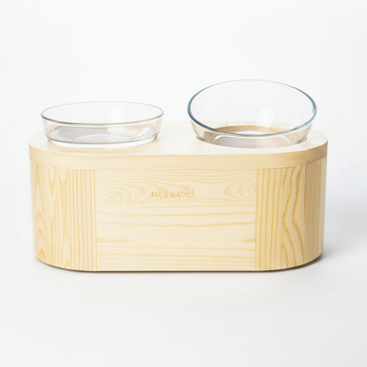 Un support de gamelle double surélevé pour chat en bois clair naturel au design minimaliste avec deux bols en verre trempé transparent, présenté sur un fond blanc, collaboration Wakupet x Palais Félin Paris.