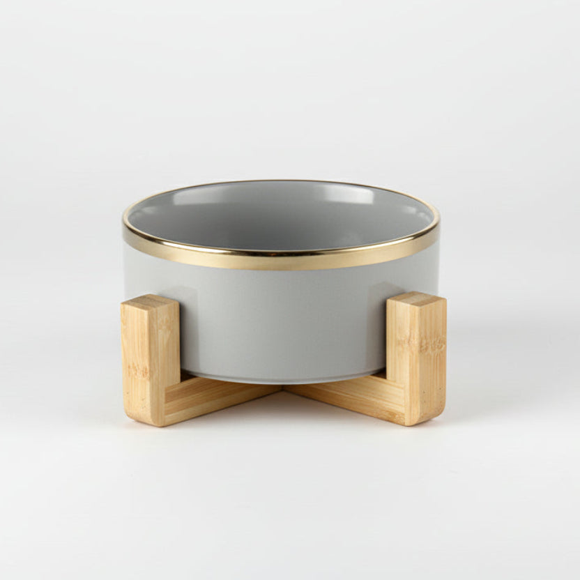 ​Gamelle individuelle haut de gamme pour chat en céramique blanche brillante avec un liseré doré élégant, posée sur un support minimaliste en bois clair croisé, signée Palais Félin Paris.