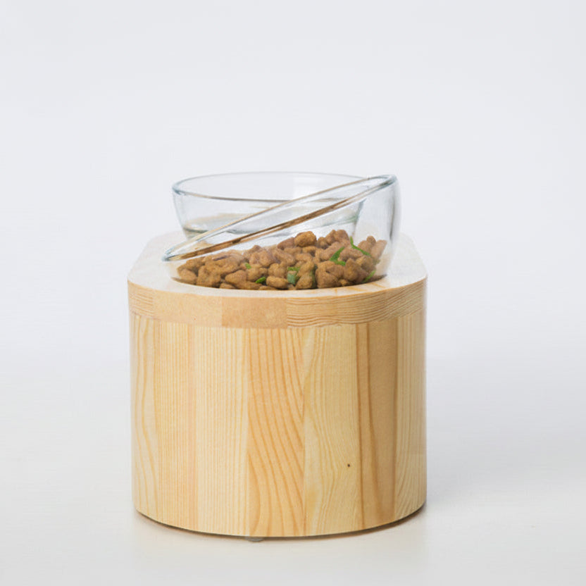 ​Une vue latérale nette, sur fond blanc uni, d'une gamelle surélevé pour chat de la marque Wakupet, issue de la collaboration avec Palais Félin Paris. L'ensemble se compose d'un support cylindrique en bois naturel clair, surmonté d'un bol en verre trempé incliné contenant des croquettes. Palais Félin Paris.
